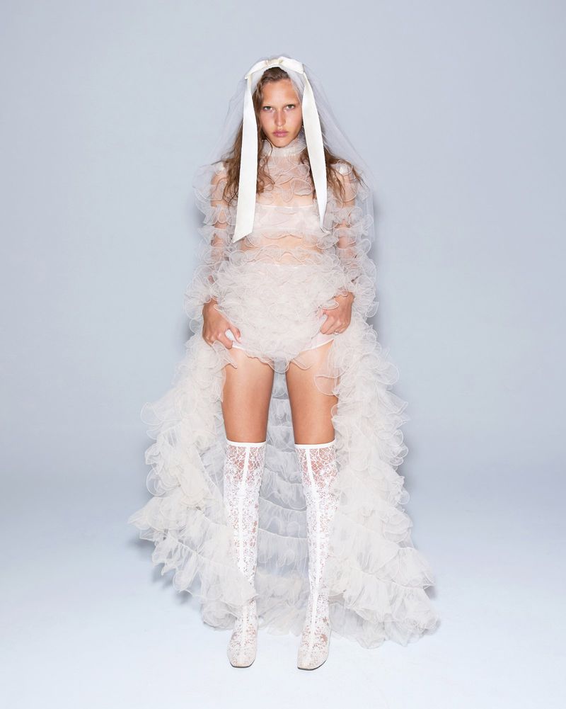 Fashion model poses in ruffled white gown with lace boots, under a veil and headpiece, in a studio setting with a pale background.