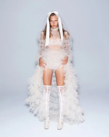 Fashion model poses in ruffled white gown with lace boots, under a veil and headpiece, in a studio setting with a pale background.