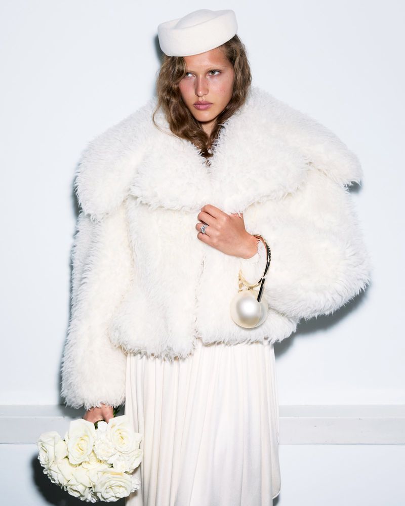 Model Angelina Kendall in an oversized white faux fur jacket and pillbox hat holding a bouquet of white roses and a pearl handbag.