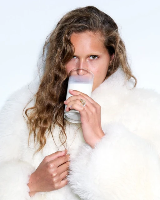 Woman in a white fur coat drinking from a glass of milk in front of a white background in a medium studio shot with natural light.