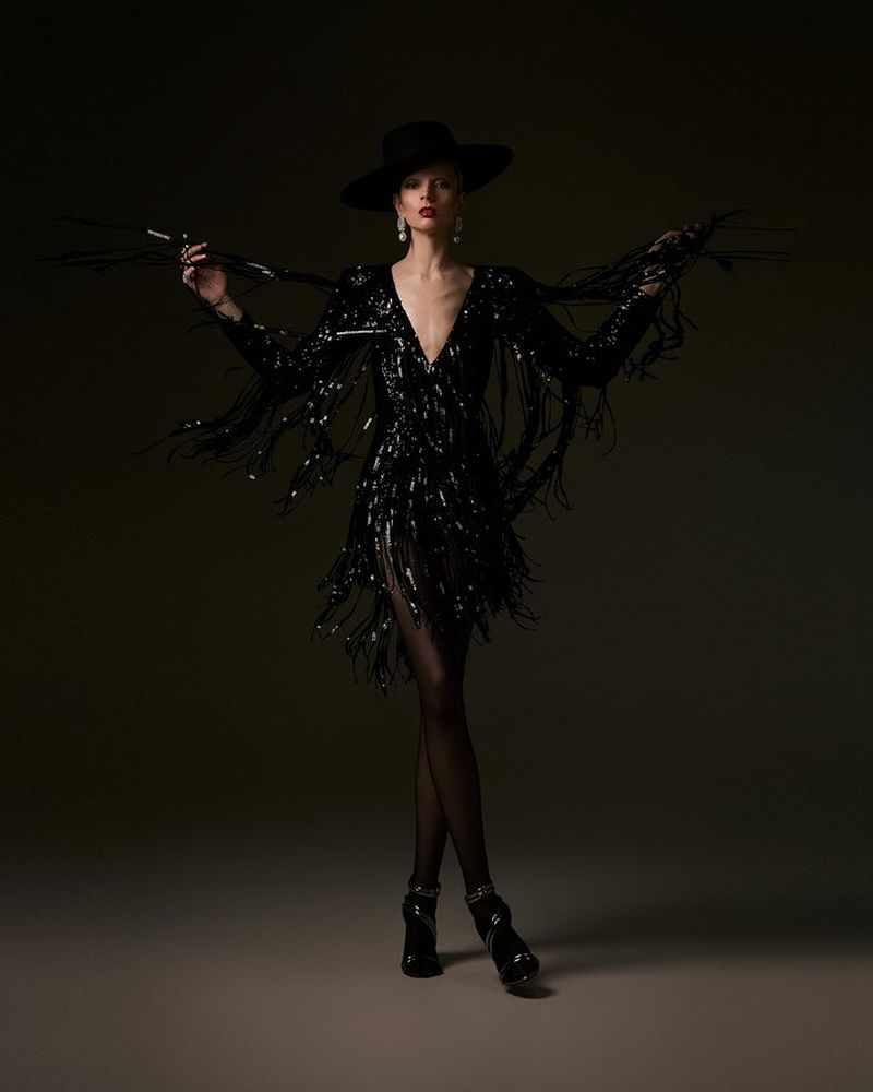 Elegant woman in a black sequined dress and wide-brimmed hat strikes a pose against a dark backdrop in a full-length studio shot.