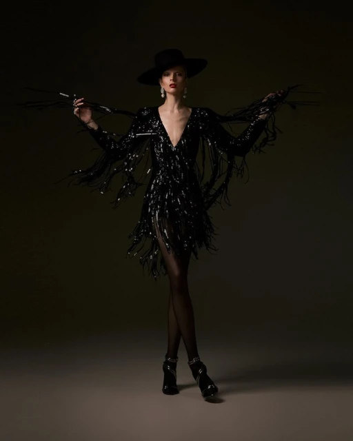 Elegant woman in a black sequined dress and wide-brimmed hat strikes a pose against a dark backdrop in a full-length studio shot.
