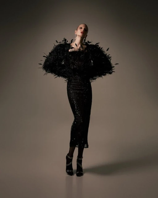 A stylish, full shot of a poised woman in a shimmering black dress and feathered bolero, complemented by strappy heels, against a neutral backdrop.