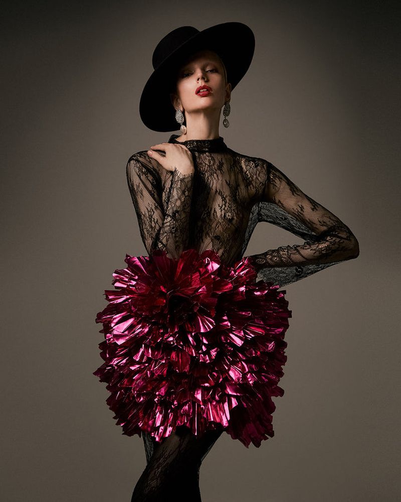A fair-skinned woman strikes a pose in a black lace top and large black hat, complemented by a red metallic flower-like skirt.