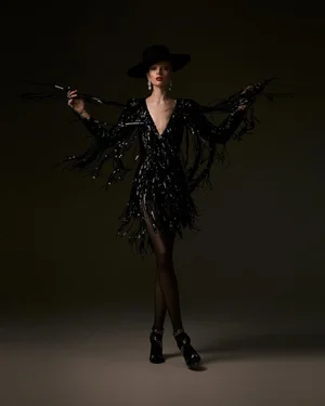 Model in black fringe sequin dress and wide-brim hat holding feathered sticks against a dark studio background