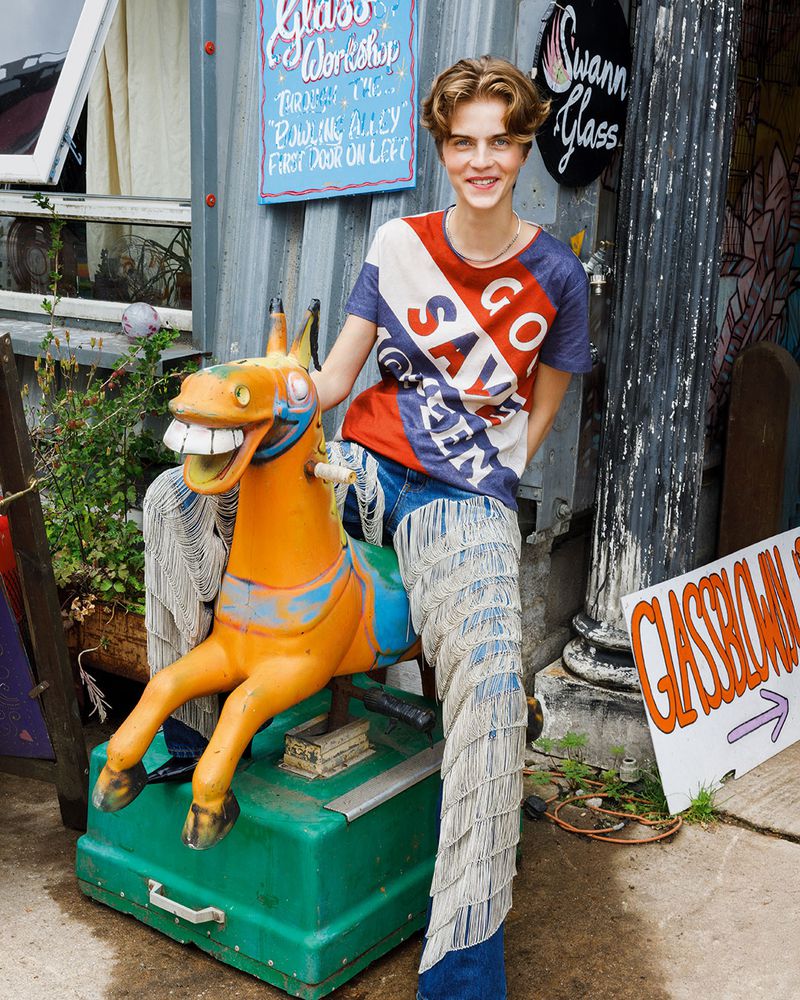 Young adult poses with a rocking horse, wearing a patterned shirt and fringed jeans outside a glass workshop in an eye-level shot.