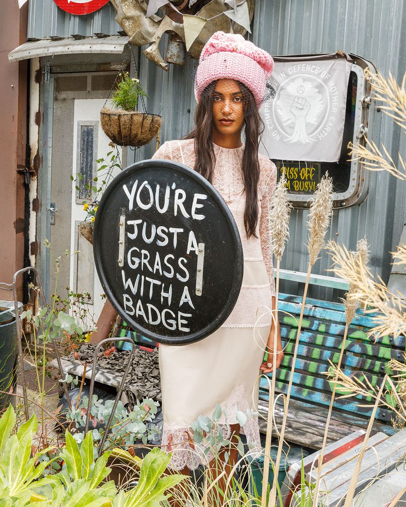 A woman wearing a pink knit hat and slip dress poses in front of a metal structure, holding a sign, surrounded by green vegetation.