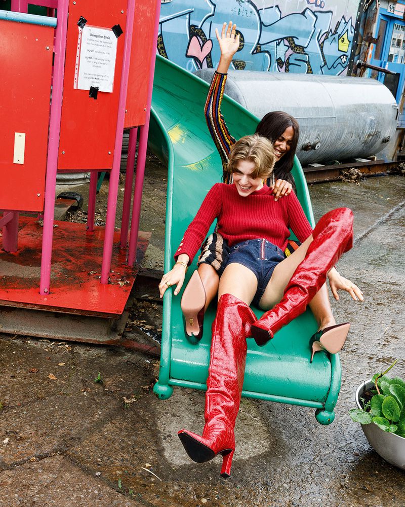 Two young women smile on a green slide with a graffiti backdrop. One wears tall red boots and a red sweater; the other raises her arm.