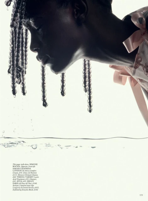 Fashion editorial shot: a black model with beaded braids, wearing a pink dress with bows, dips her face in water. Studio lighting.