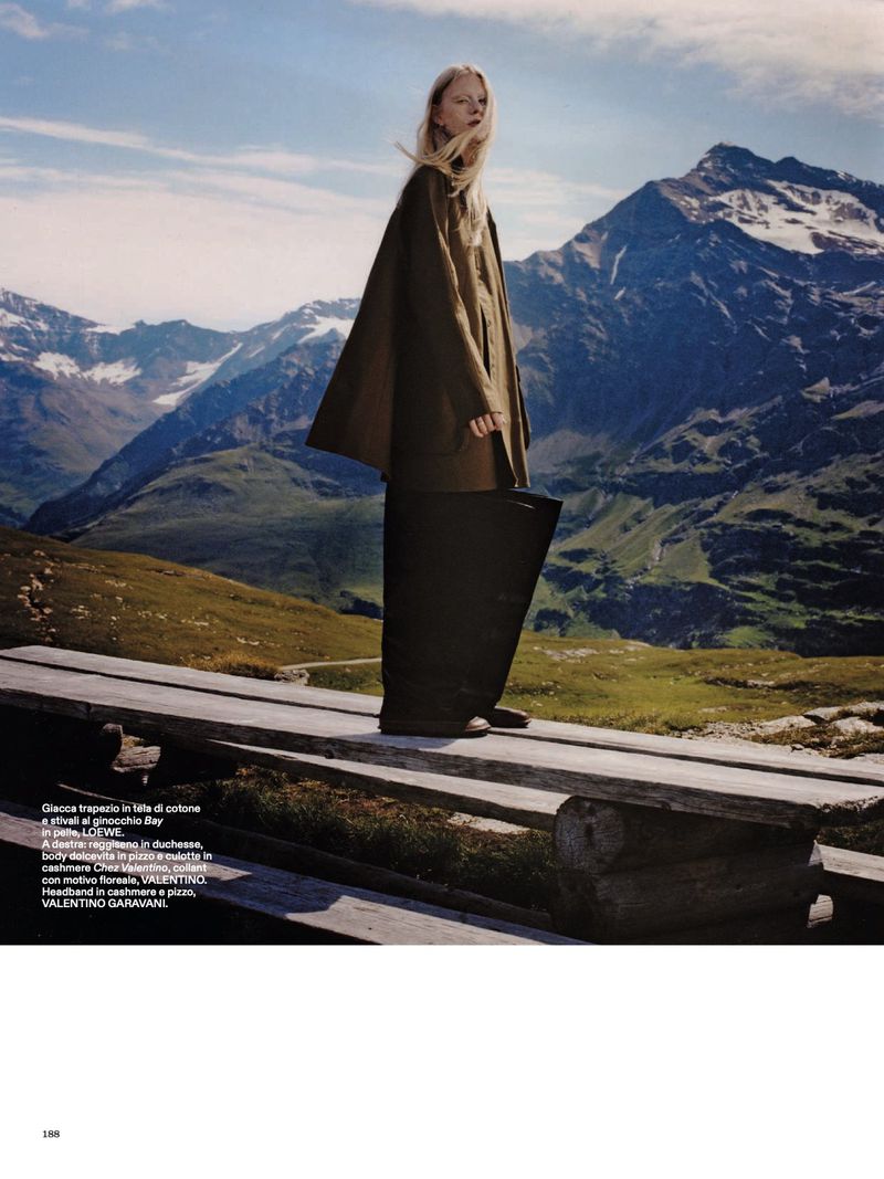 A blonde model poses on a wooden path in the mountains, wearing a brown cape and holding a black bag against a mountain backdrop.