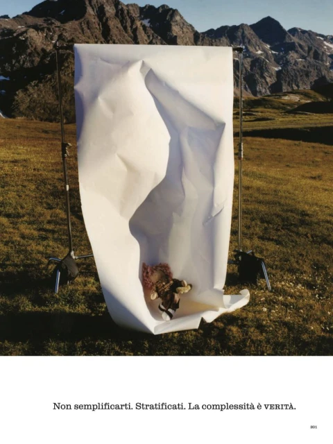 Toy animal under a large crumpled sheet of white paper propped up by stands in a grassy field against a mountain backdrop.