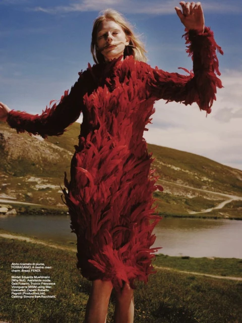 Fashion editorial shot of a woman in a red feathered dress standing on a hillside with a lake and mountains in the background.