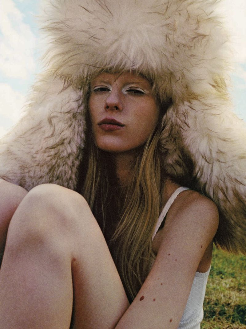Model Eduarda Muehlmann wearing an oversized cream faux fur hat and white tank top seated outdoors against an open sky.