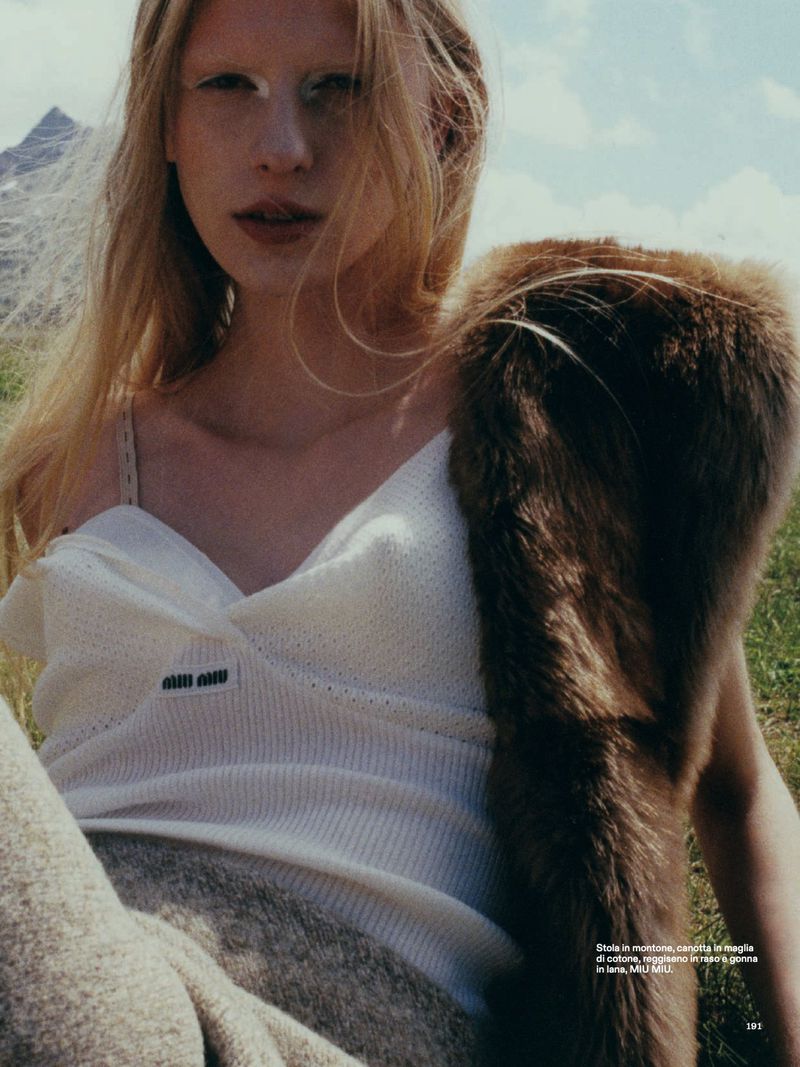 Fashion editorial shot of a blonde woman wearing a white top and fur stole, posing outdoors against a blurred mountain backdrop.