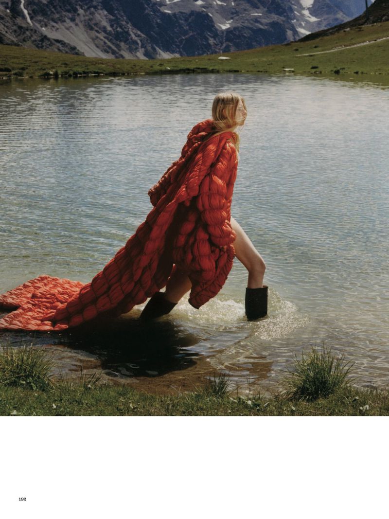 Woman in a red puffer coat and boots stands in a lake with mountains in the background, creating a fashion editorial shot.