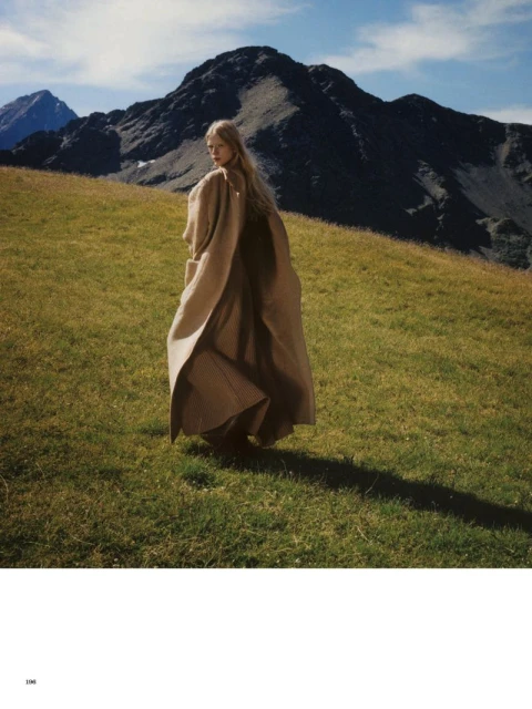 A lone female figure wearing a flowing tan coat stands on a grassy hill with mountains rising in the background under a blue sky.