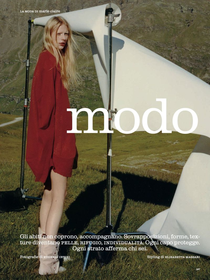 Editorial fashion shot of a blonde model in a red oversized sweater posing in an outdoor setting with a white reflector and mountain backdrop.