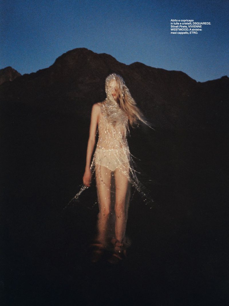 A slender adult female stands in water, adorned in a sparkling, fringed garment, against a mountain backdrop under a twilight sky.
