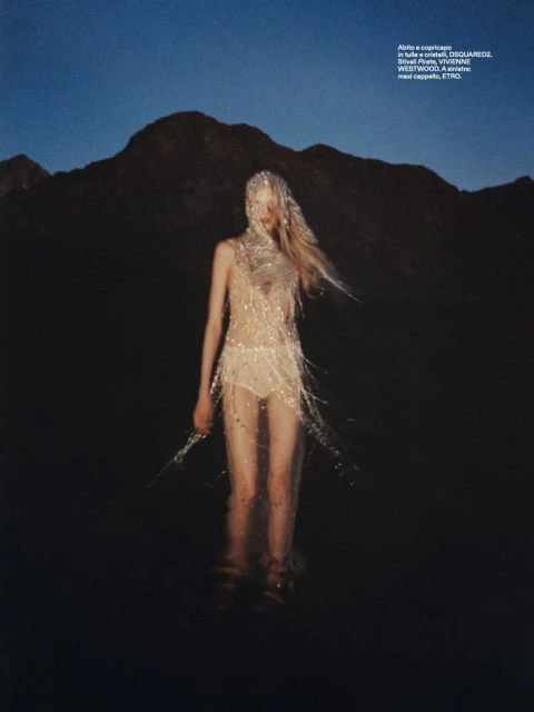 A slender adult female stands in water, adorned in a sparkling, fringed garment, against a mountain backdrop under a twilight sky.