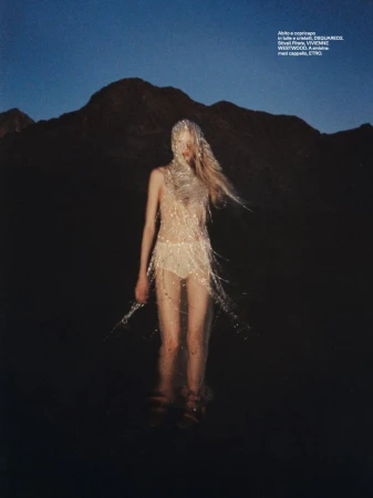 A slender adult female stands in water, adorned in a sparkling, fringed garment, against a mountain backdrop under a twilight sky.
