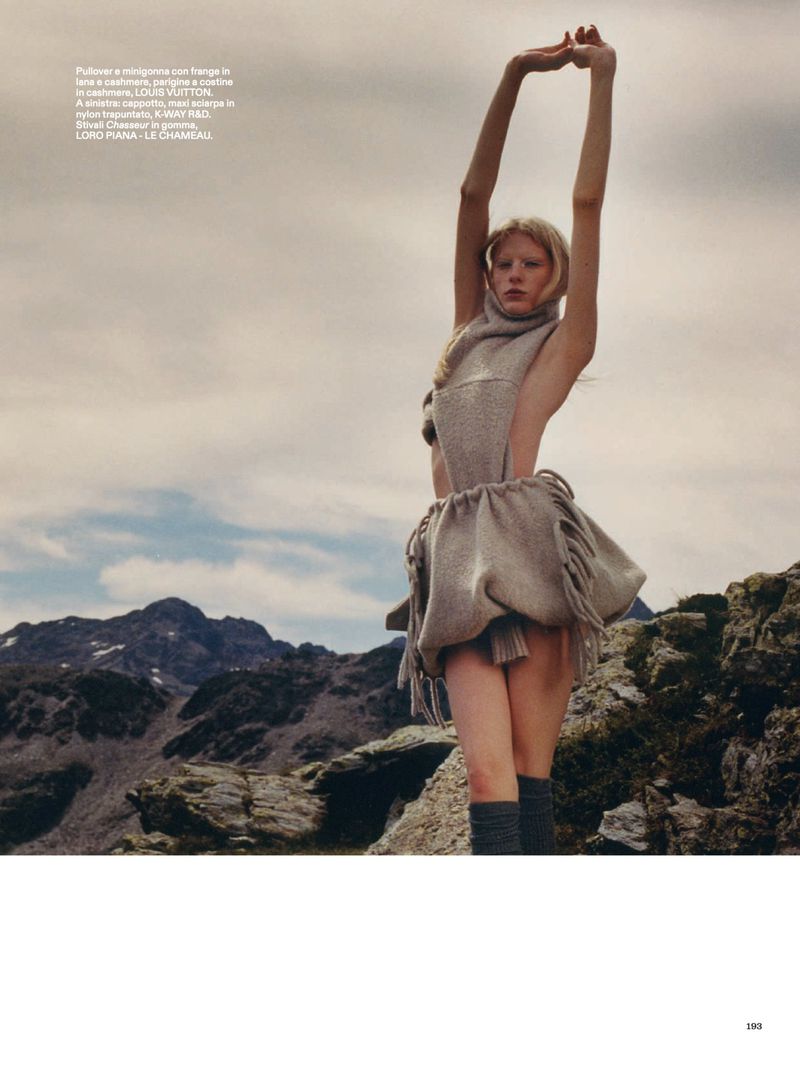 Woman in fringed dress stands with arms raised in a mountain landscape under a cloudy sky for a fashion editorial outdoors.