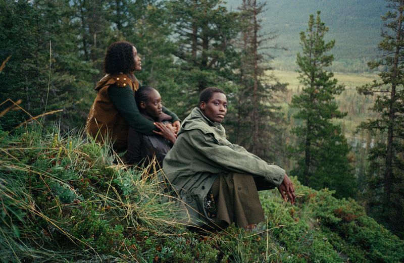 Three women sit on a grassy hillside, gazing out over a forest and valley landscape in daylight. They are wearing earthy-toned clothing.