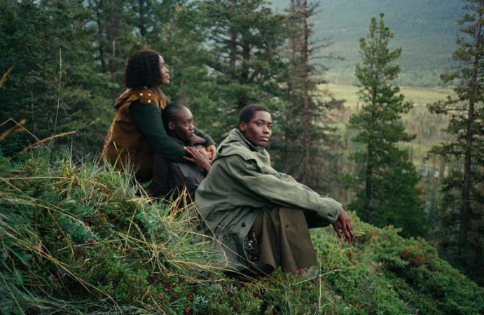 Three women sit on a grassy hillside, gazing out over a forest and valley landscape in daylight. They are wearing earthy-toned clothing.