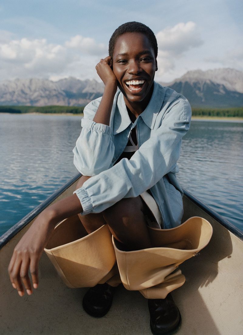 Smiling African woman in a canoe on a lake with distant mountains. She wears a light blue jacket, tan pants, and black shoes.