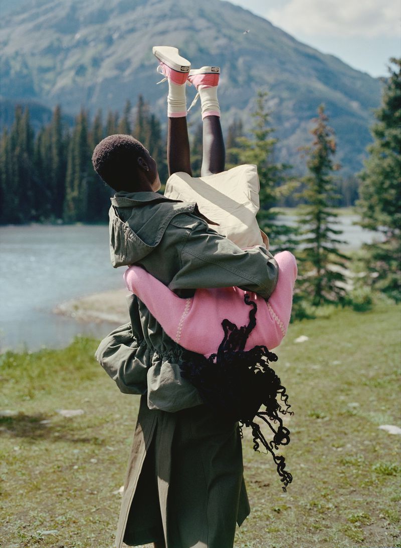 Two people in an outdoor setting, one is being lifted upside down by the other, set against a scenic mountain and lake backdrop.