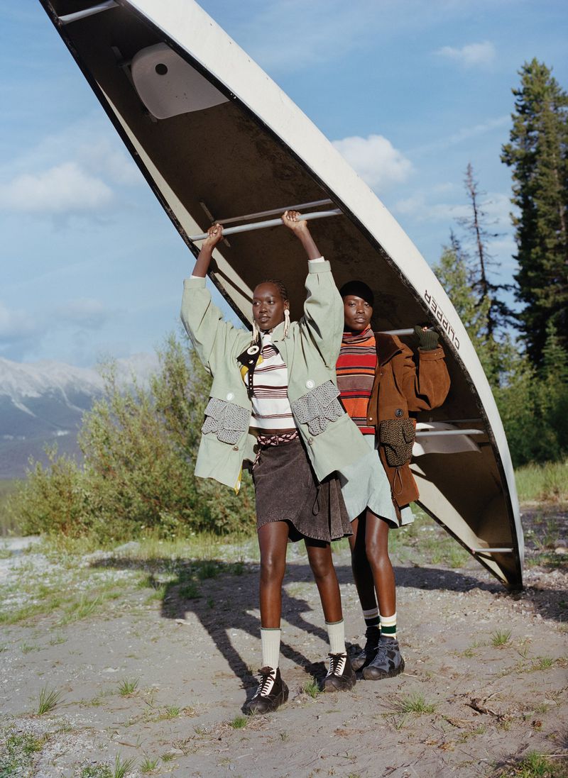 Two women in fashionable outfits carry a canoe. They are outdoors, with a grassy area beneath their feet and mountain scenery in the distance.