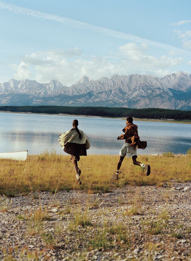 Two models in outdoor fashion run toward an alpine lake with rugged mountain peaks in the background for American Vogue.