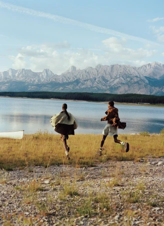 Two models in outdoor fashion run toward an alpine lake with rugged mountain peaks in the background for American Vogue.
