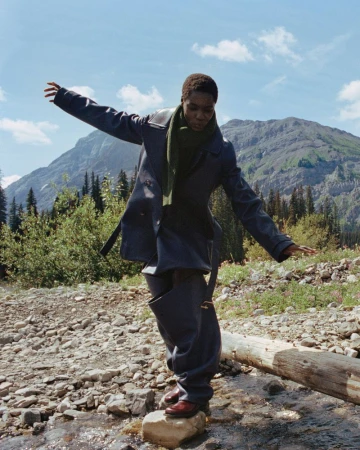 Model Awar Odhiang balancing on a rock in a mountain stream wearing an oversized navy leather coat and olive scarf.