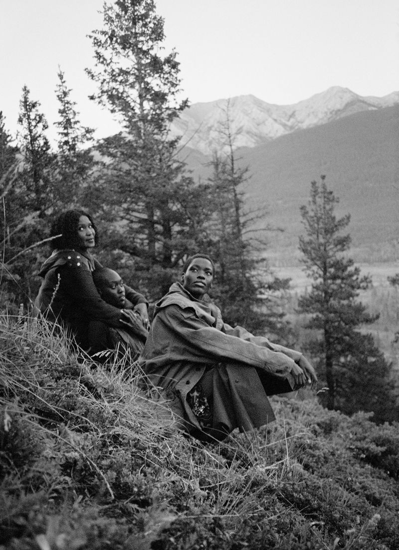 Three people, two females and one male, sit on a grassy hillside with a mountain range in the background in a black and white outdoor shot.