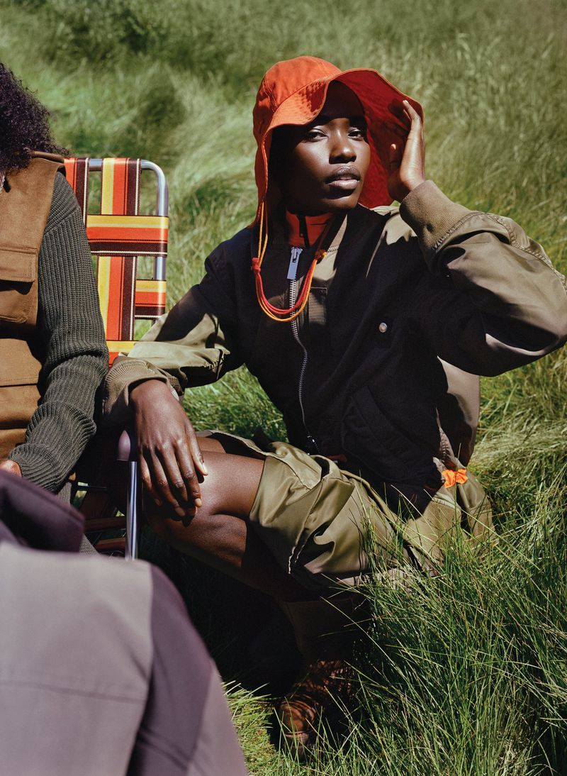 Two dark-skinned women sitting in a field of tall green grass, wearing outdoor utility clothing with orange hats and trim.