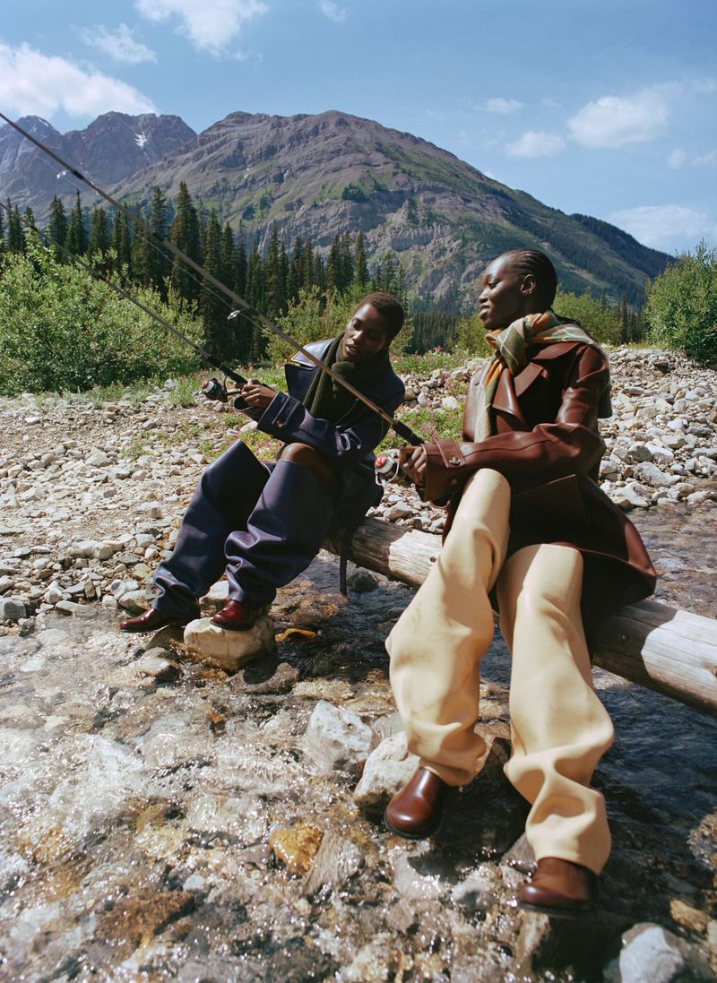 Two people in stylish outdoor clothing are fishing near a mountain stream with a backdrop of mountains and trees on a sunny day.