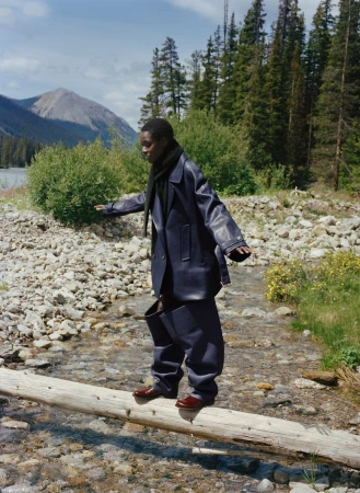 A dark-skinned man in an oversized blue suit carefully balances on a fallen log over a stream, surrounded by trees and mountains.