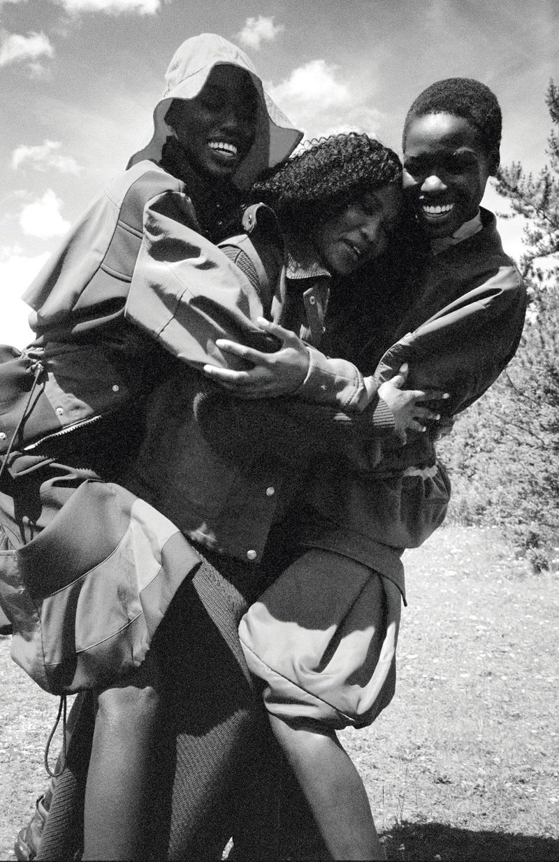 Three smiling people embrace outside in a black and white photograph. Two are dark-skinned, and the third person in the middle is lighter-skinned.