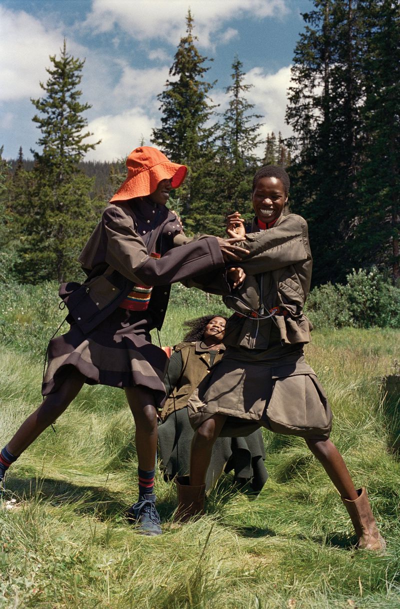 Three African models pose in a grassy field wearing retro-inspired fall fashion, featuring skirts, hats, jackets, and boots.