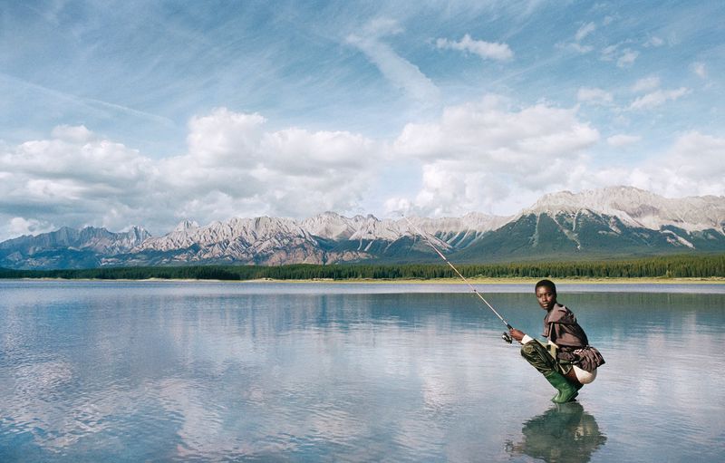 Person fishing in still lake with backdrop of tree line and cloud-covered mountains, reflected in water with sky and clouds above.