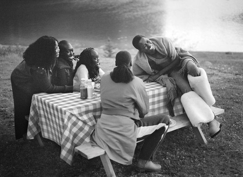 Five women sitting at a picnic table in a black and white shot. One woman is laughing with her feet up. A picnic blanket covers the table.