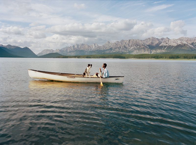 An adult and a dog are in a canoe on a lake with mountains in the background under a partly cloudy sky, capturing a serene outdoor moment.