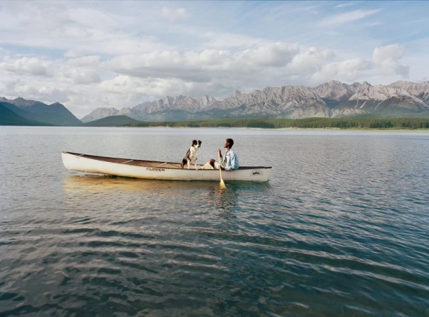 An adult and a dog are in a canoe on a lake with mountains in the background under a partly cloudy sky, capturing a serene outdoor moment.