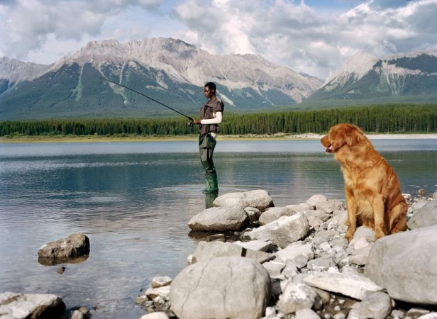 An adult fishes in a lake with a dog on the rocky shore, set against a background of trees and mountains under a cloudy sky.