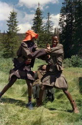 Models in outerwear posing dynamically in a grassy clearing with tall pine trees under an overcast sky.