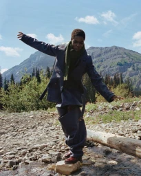Model Awar Odhiang balancing on a rock in a mountain stream wearing an oversized navy leather coat and olive scarf.
