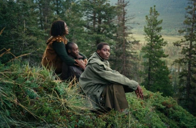 Three Black women dressed in oversized olive and earthy outerwear sit among pine trees on a grassy hillside.