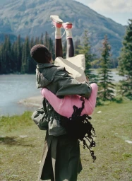 A person in an olive green parka carries model Awar Odhiang upside down in a pink sweater against an alpine mountain backdrop.