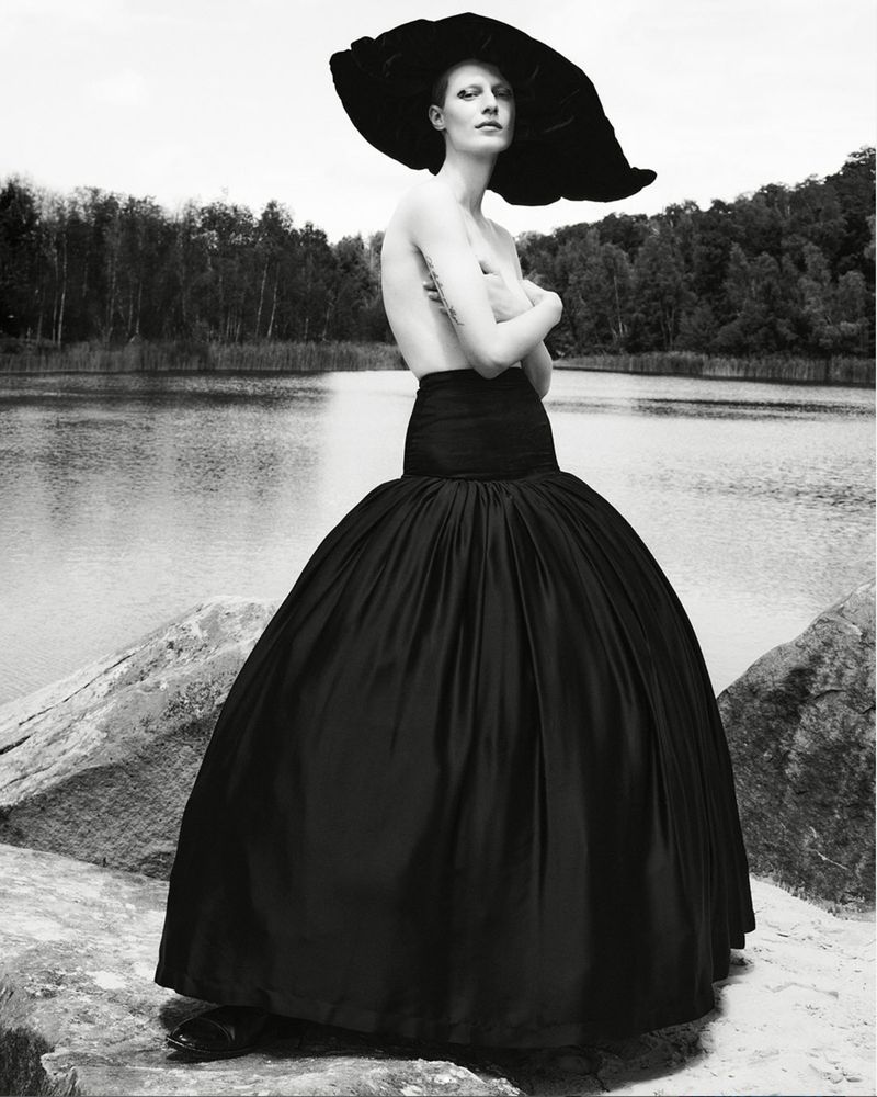 Monochrome portrait of a woman in a large hat and voluminous skirt posing outdoors by a lake surrounded by trees.