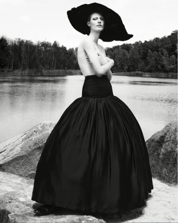 Monochrome portrait of a woman in a large hat and voluminous skirt posing outdoors by a lake surrounded by trees.